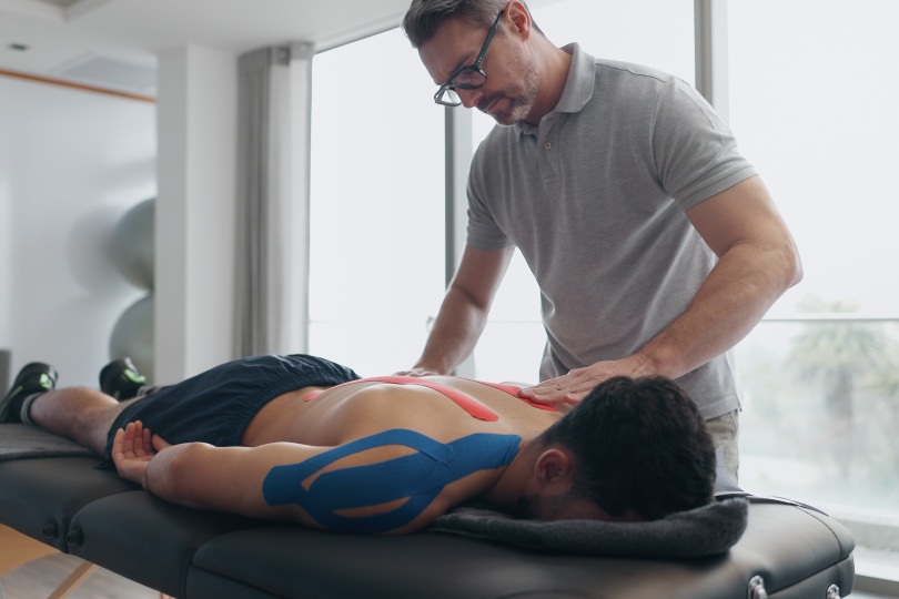 Fisioterapeuta, persona y cinta en cama para lesión con proceso de recuperación, dolor de espalda y ayuda en clínica. Persona, biocinética y tratamiento para rehabilitación con músculo, columna y servicio en hospital