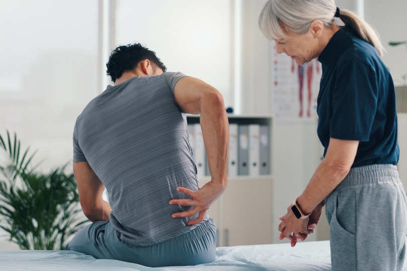 Dolor de espalda, fisioterapia y paciente en consulta por problema, inflamación o distensión muscular. Personas, quiropráctico y tratamiento para lesiones, rehabilitación y recuperación de ligamentos de la columna vertebral.