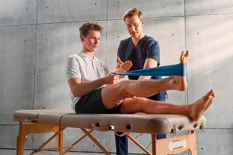 Sport Physiotherapy Specialist Showing How to Stretch a Rubber Band on Specific Muscle Groups or Joints to Young Male Athlete. Sportsman Recovering from Mild Injury, Undergoing Rehabilitation.